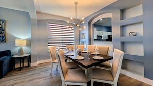 Un coin repas avec une table en bois foncé, dressée pour six personnes, entourée de chaises blanches, et un lustre moderne au-dessus.