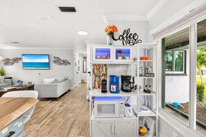 A modern kitchen with a coffee station, a dining area with a table and chairs, a living room with a sofa and a TV, and a large window with a view of greenery. - TIKI on the CANAL mini-putt pool/spa kayak fishing (Cape Coral)