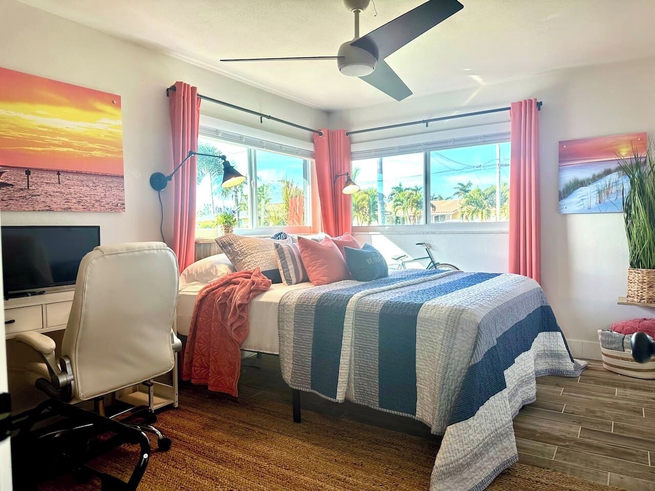 A bedroom with a bed, a desk, a chair, a TV, and large windows with curtains.
