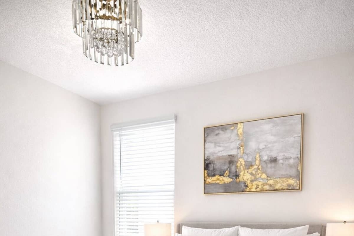 A bedroom with a bed, bedside tables, a chandelier, a window with blinds, and a painting on the wall.