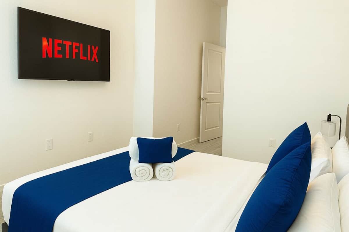 A hotel room with a bed, a wall-mounted TV displaying the Netflix logo, a door, and a light fixture.