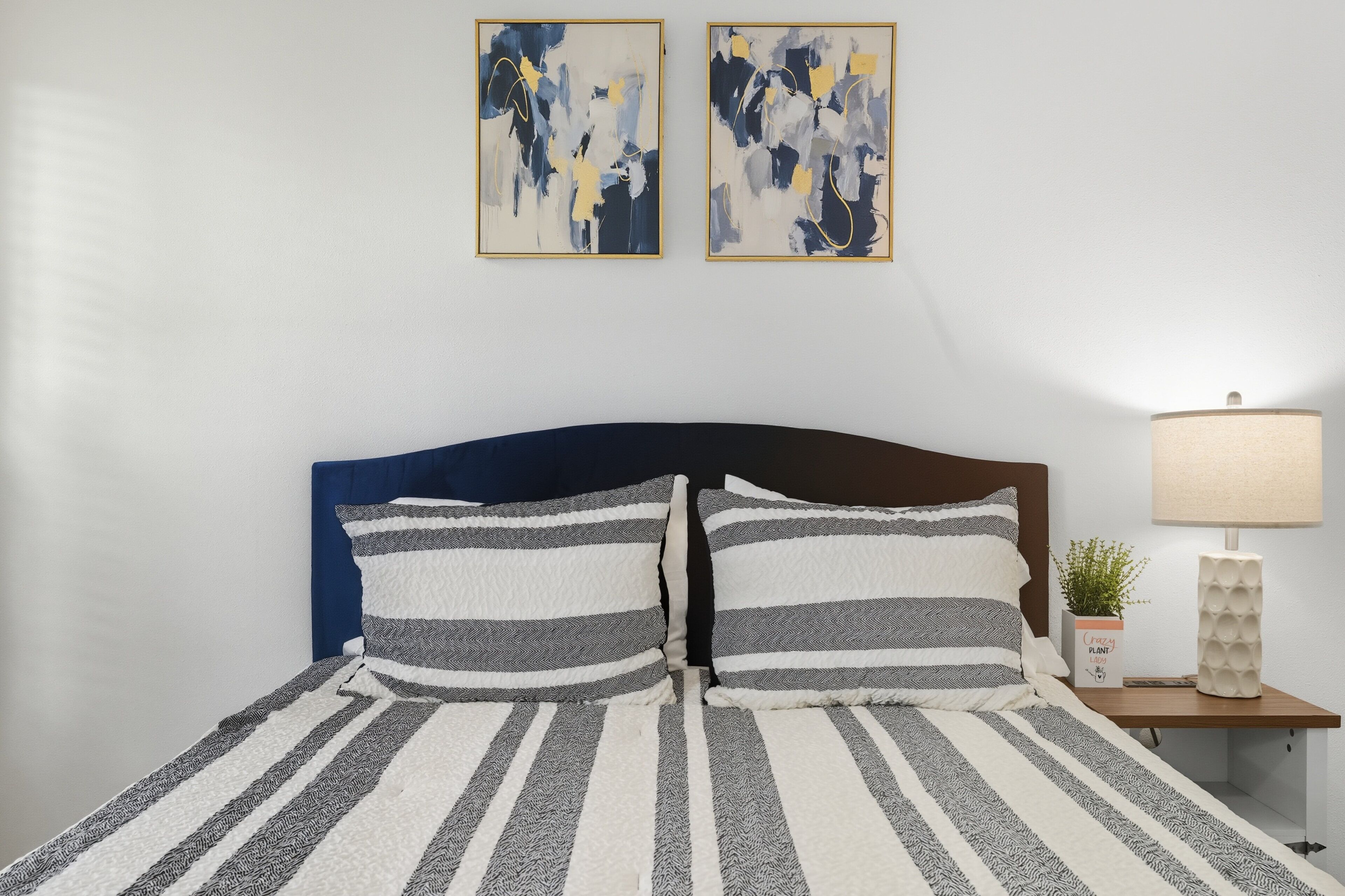 A bed with striped bedding, a nightstand with a lamp and a plant, and abstract art on the wall.
