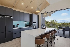 A modern kitchen with dark cabinets, a white island, and bar stools. - Panorama Lookout | Brand New Spacious Winter Retreat with Ocean Views (Normanville)