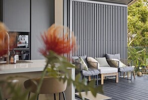 A modern outdoor seating area with a wooden deck, a metal railing, and a vertical striped wall in the background. - Panorama Lookout | Brand New Spacious Winter Retreat with Ocean Views (Normanville)