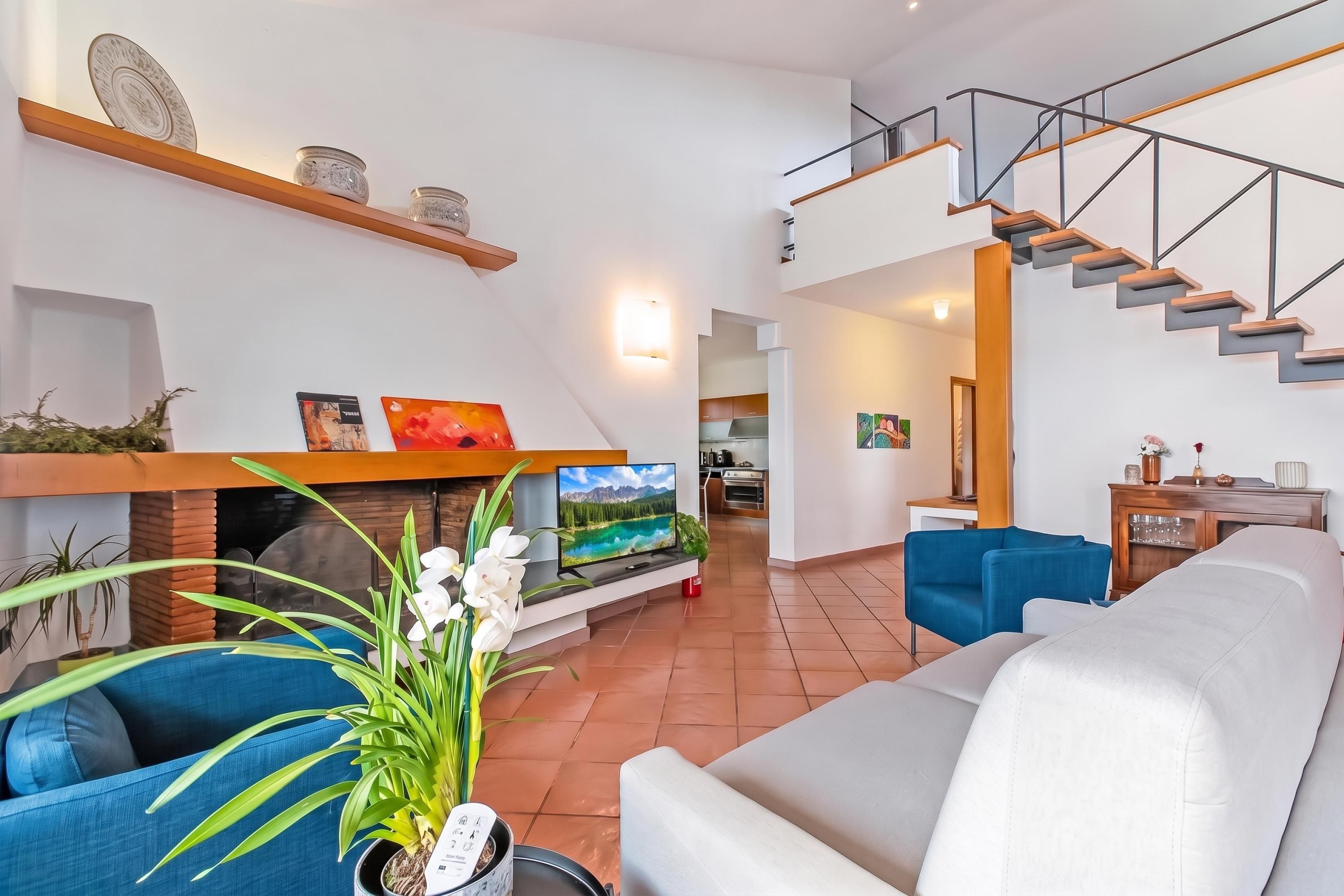 Een moderne woonkamer met een bank, een televisie en een open haard. Er is een trap die naar een hoger gelegen verdieping leidt.