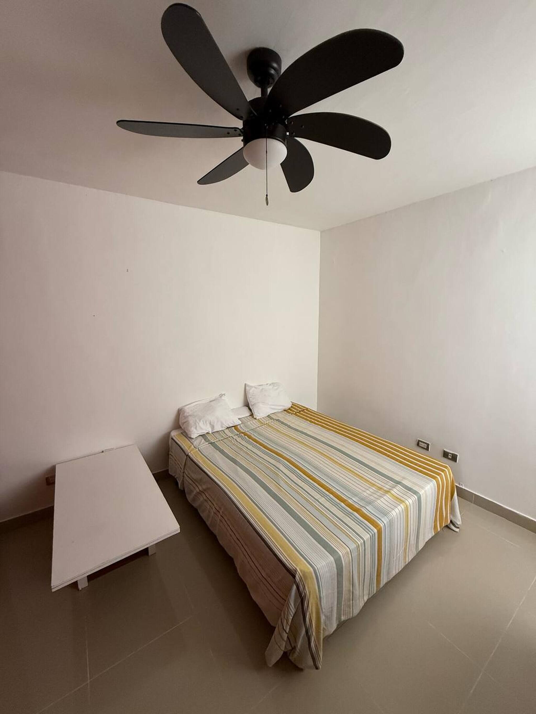 Une chambre à coucher avec un lit, une table de chevet blanche, un ventilateur de plafond et un mur uni.