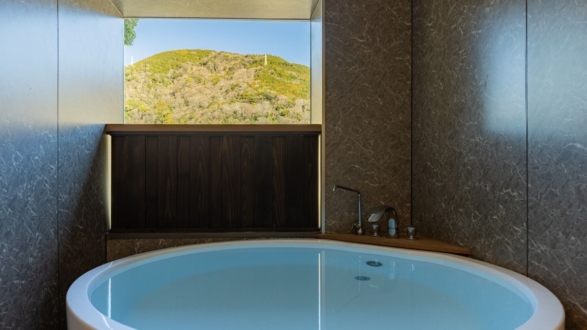 A modern bathroom with a large bathtub, a wooden vanity, and a view of a hilly landscape.