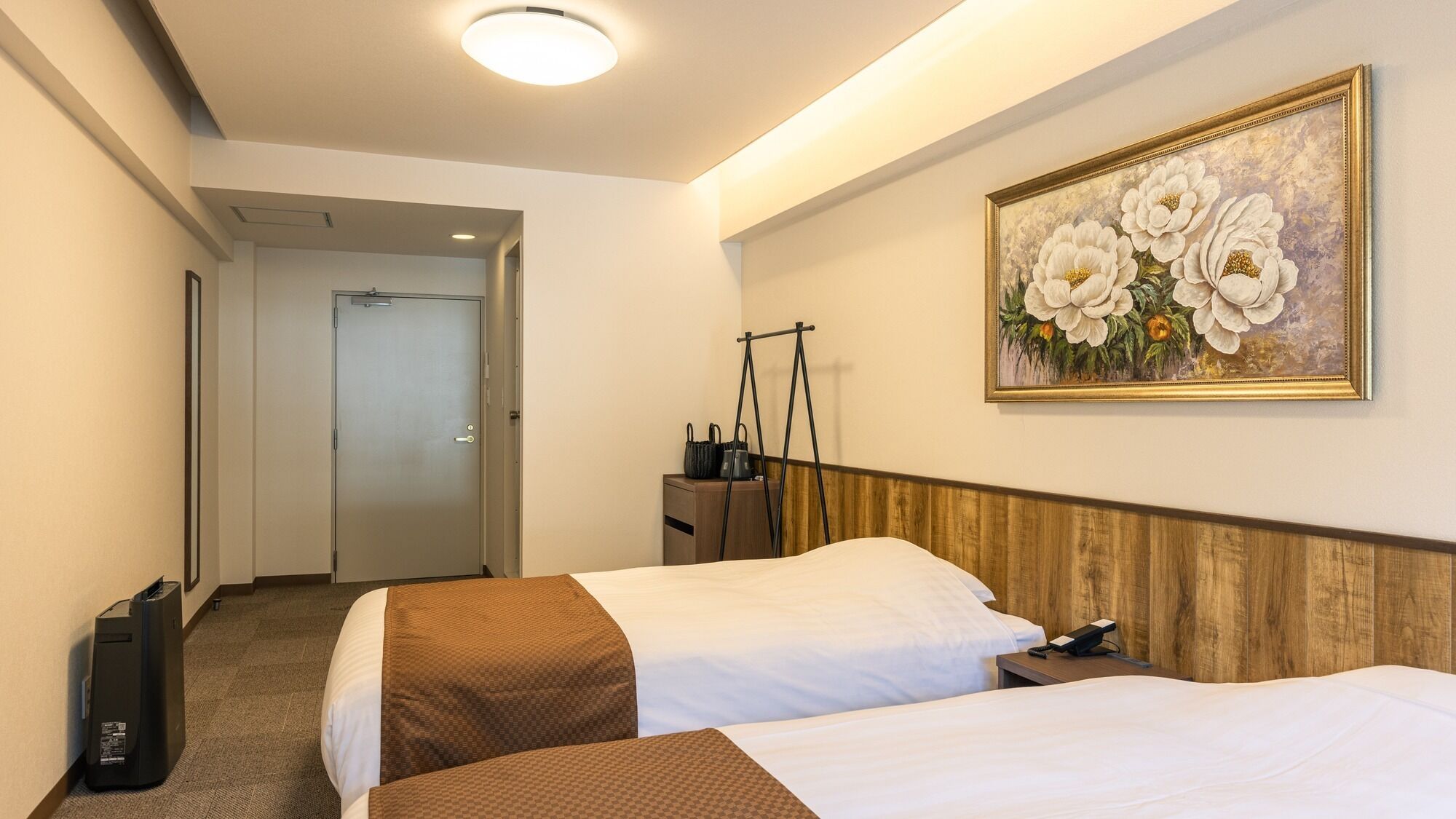 A hotel room with two beds, a painting of flowers, a desk with a lamp, and a suitcase.