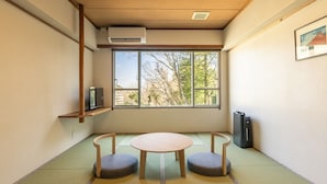 Une pièce dotée d’un sol recouvert de tatamis, d’une table en tatami, de deux bancs en tatami, d’un climatiseur fixé au mur, d’une étagère avec un téléviseur et d’une fenêtre donnant sur des arbres et des bâtiments.