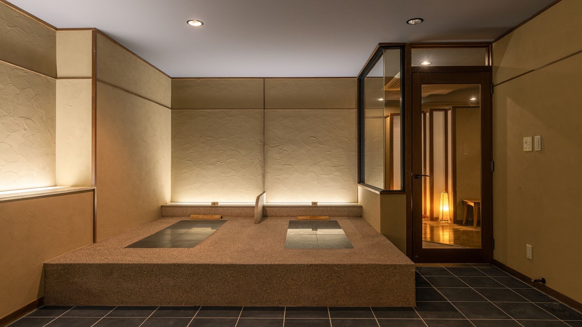 A room with a large, rectangular, beige-tiled floor, a small window, and a door leading to another room.