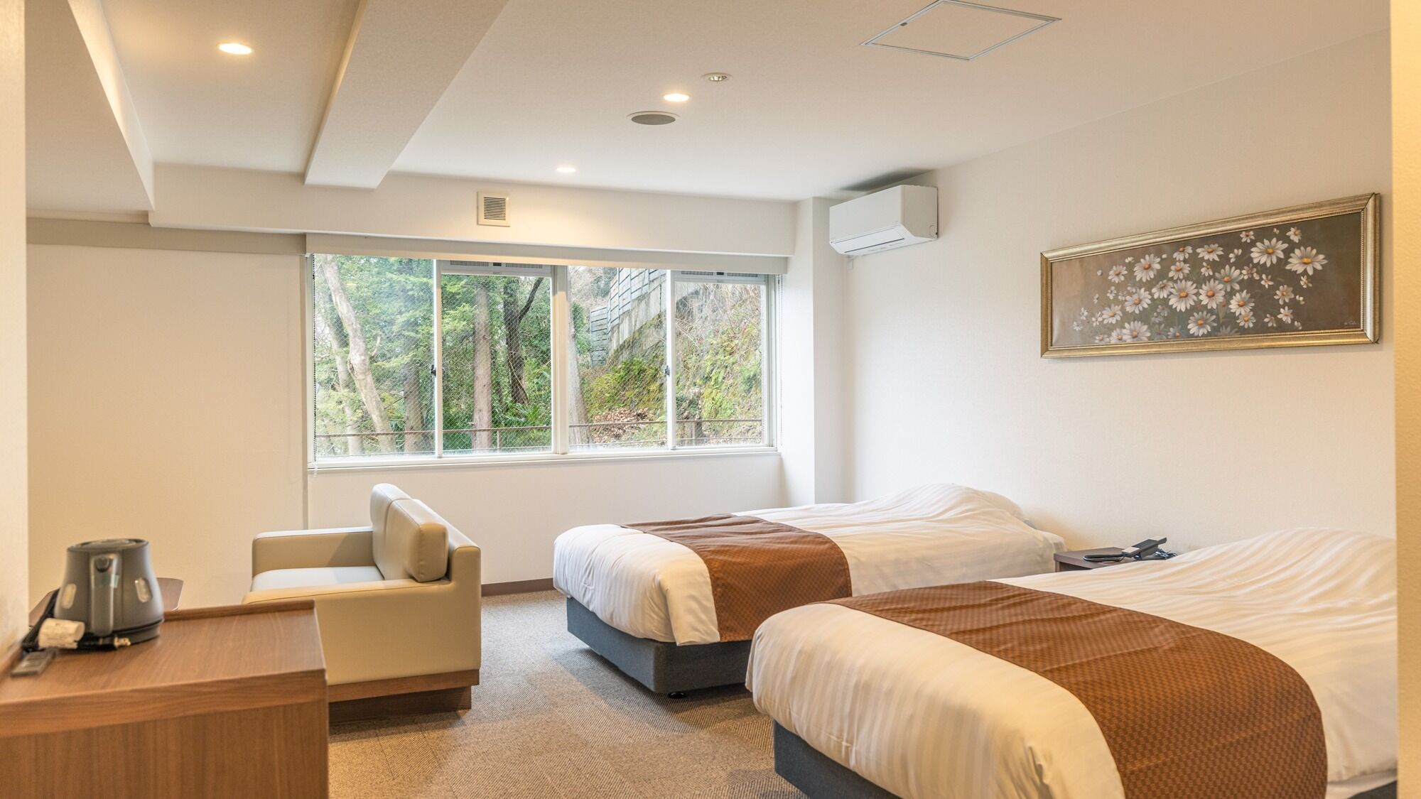 A hotel room with two beds, a desk, a chair, and a large window with a view of trees.