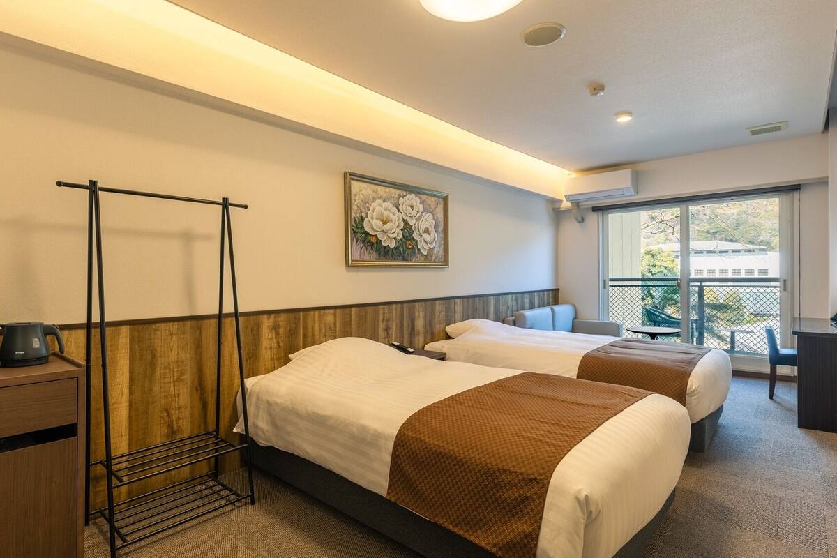 Une chambre d’hôtel avec deux lits, une tête de lit en bois, un tableau, un balcon avec vue et un bureau avec une bouilloire.