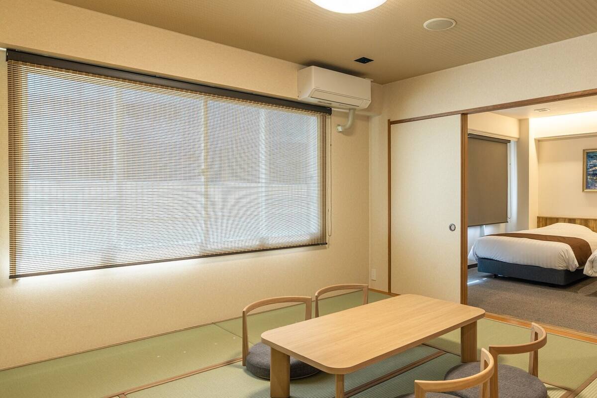 Une chambre d’hôtel avec un sol recouvert de tatamis, une table en bois avec des chaises, une grande fenêtre avec des stores et un tableau accroché au mur.