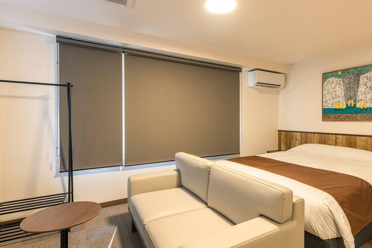 Une chambre d’hôtel comprenant un lit, un canapé, une petite table, une fenêtre avec des stores, un appareil de climatisation et un tableau au mur.
