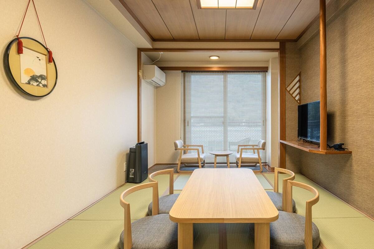 Une pièce avec une table en bois, quatre chaises, un téléviseur posé sur une étagère et une grande fenêtre avec des stores.