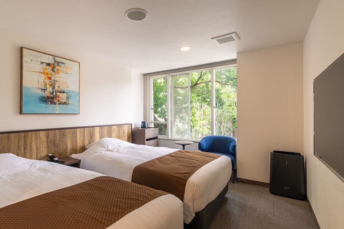 Une chambre d’hôtel avec deux lits, un tableau, une fenêtre donnant sur des arbres, un bureau et une chaise.