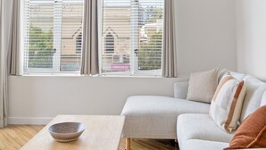 A modern living room with a sofa, a wooden table, and a view through large windows. - The Lookout – Modern Fremantle Apartment with View (Fremantle)
