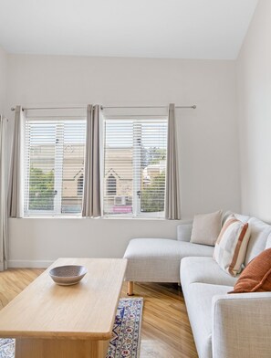 A modern living room with a sofa, a wooden table, and a view through large windows. - The Lookout – Modern Fremantle Apartment with View (Fremantle)