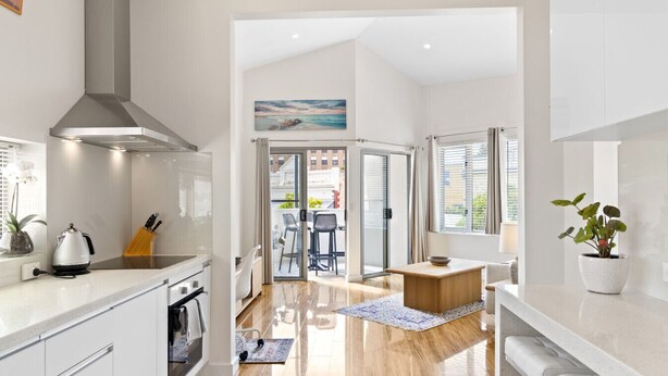 A modern kitchen with a stove, sink, and overhead ventilation. There is a dining area with a table and chairs, and a view through a glass door to an outdoor space. - The Lookout – Modern Fremantle Apartment with View (Fremantle)