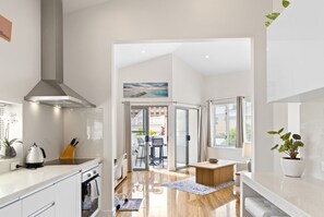 A modern kitchen with a stove, sink, and overhead ventilation. There is a dining area with a table and chairs, and a view through a glass door to an outdoor space. - The Lookout – Modern Fremantle Apartment with View (Fremantle)