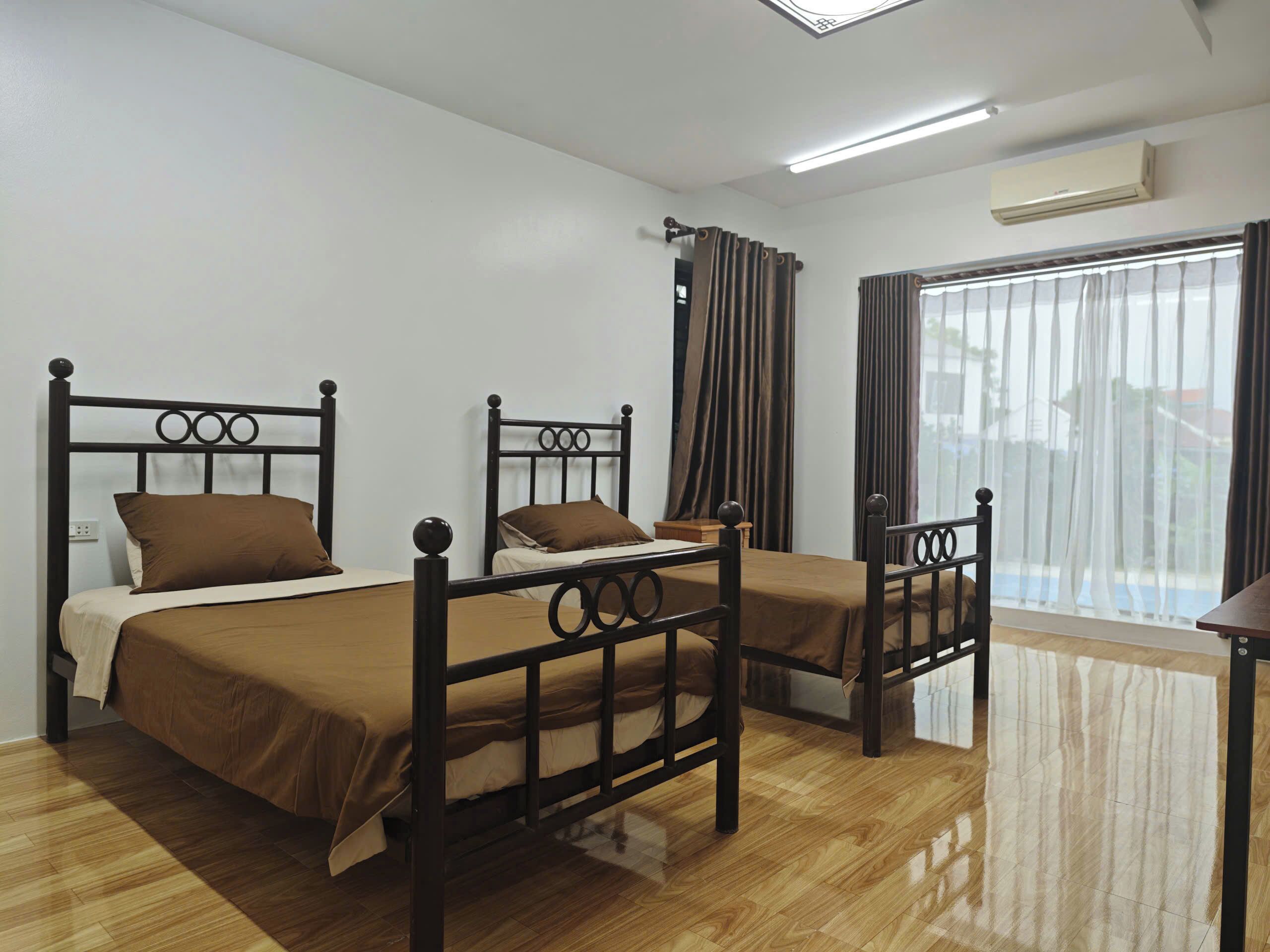 A room with two beds, a window with curtains, an air conditioning unit, and a wooden floor.