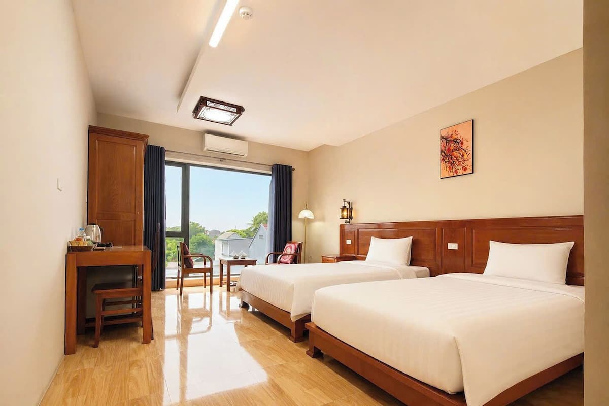 A hotel room with two beds, a desk, a chair, a lamp, and a view of the outdoors.