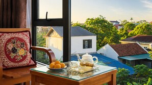 Une pièce avec vue sur des maisons, une table avec une théière et des oranges, et une chaise agrémentée d’un coussin décoratif.