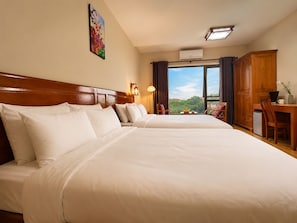 A hotel room with two beds, a desk with a laptop, a chair, a small table with fruit, and a window with a view of greenery.