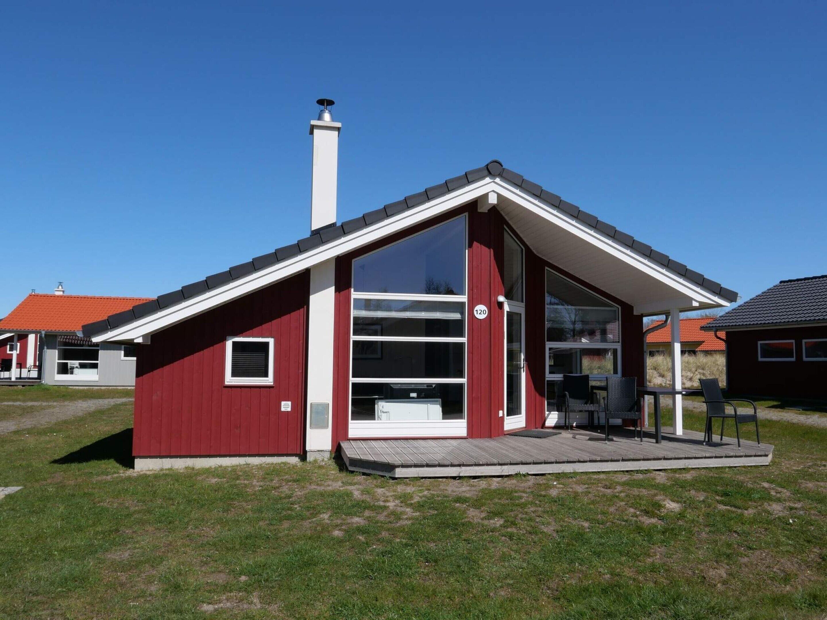 En modern, röd byggnad med ett stort glasfönster, en skorsten och en liten veranda med stolar.