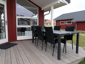 Eine überdachte Terrasse mit Tisch und Stühlen, ein Glastür-Eingang und ein Blick auf ein rotes Gebäude im Hintergrund.