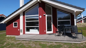 Une maison moderne avec une façade rouge, de grandes fenêtres et une terrasse aménagée avec du mobilier d’extérieur.