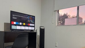 Hotellihuone, jossa on televisio, josta näkyvät eri kanavat, työpöytä, tuoli, lattialämmitin, ikkuna, josta on näkymä, ja sänky, jossa on violetti päiväpeite.