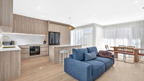 Een moderne keuken met een ingebouwde oven, een eethoek met een houten tafel en stoelen, en een blauwe bank in de woonkamer.