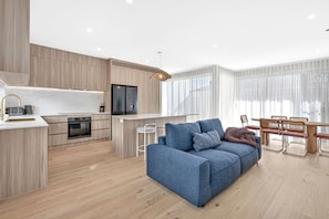 Een moderne keuken met een ingebouwde oven, een eethoek met een houten tafel en stoelen, en een blauwe bank in de woonkamer.