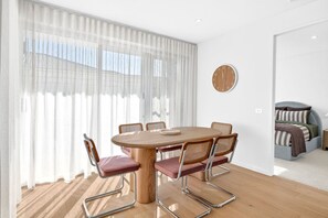 Een kamer met een ronde houten tafel, zes stoelen, een klok aan de muur en een bed met gestreept beddengoed.