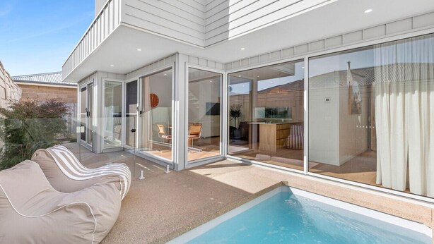 Een modern huis met een zwembad, glazen deuren die leiden naar een terras, en een zichtbare keukenruimte.