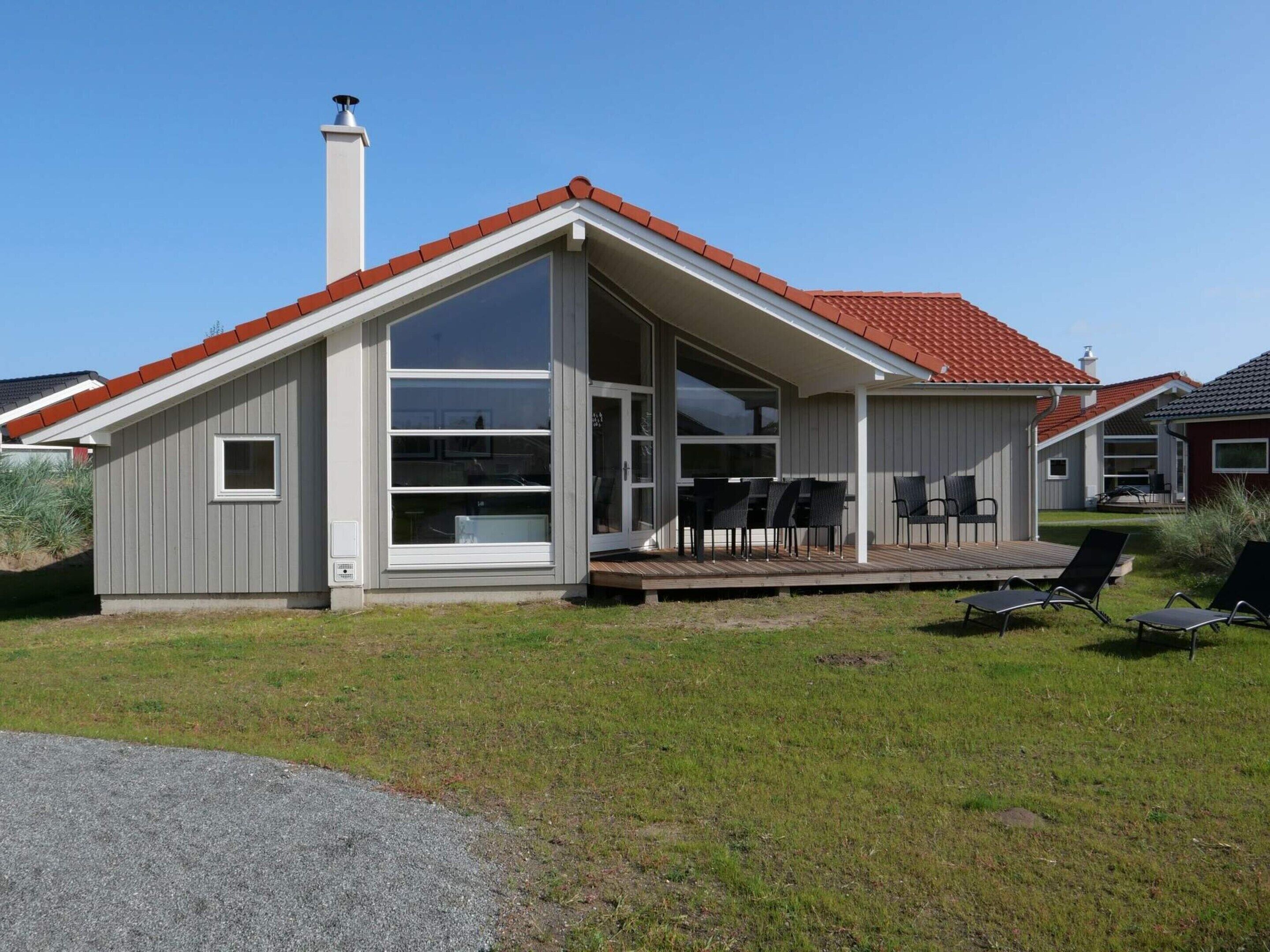 Uma casa moderna com uma grande janela de vidro, um deck com mobiliário de exterior e um telhado de telhas vermelhas.