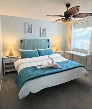 Ein ordentlich gemachtes Bett mit einer weiß-blauen Bettdecke, zwei passende Kissen, ein weißes Dekorationsaccessoire auf dem Bett, ein Deckenventilator und ein Nachttisch mit einer Lampe.