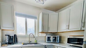 Eine moderne Küche mit hellen Schränken, einem Edelstahlkühlschrank, einer Mikrowelle, einer Kaffeemaschine und einem Fenster mit Jalousien.