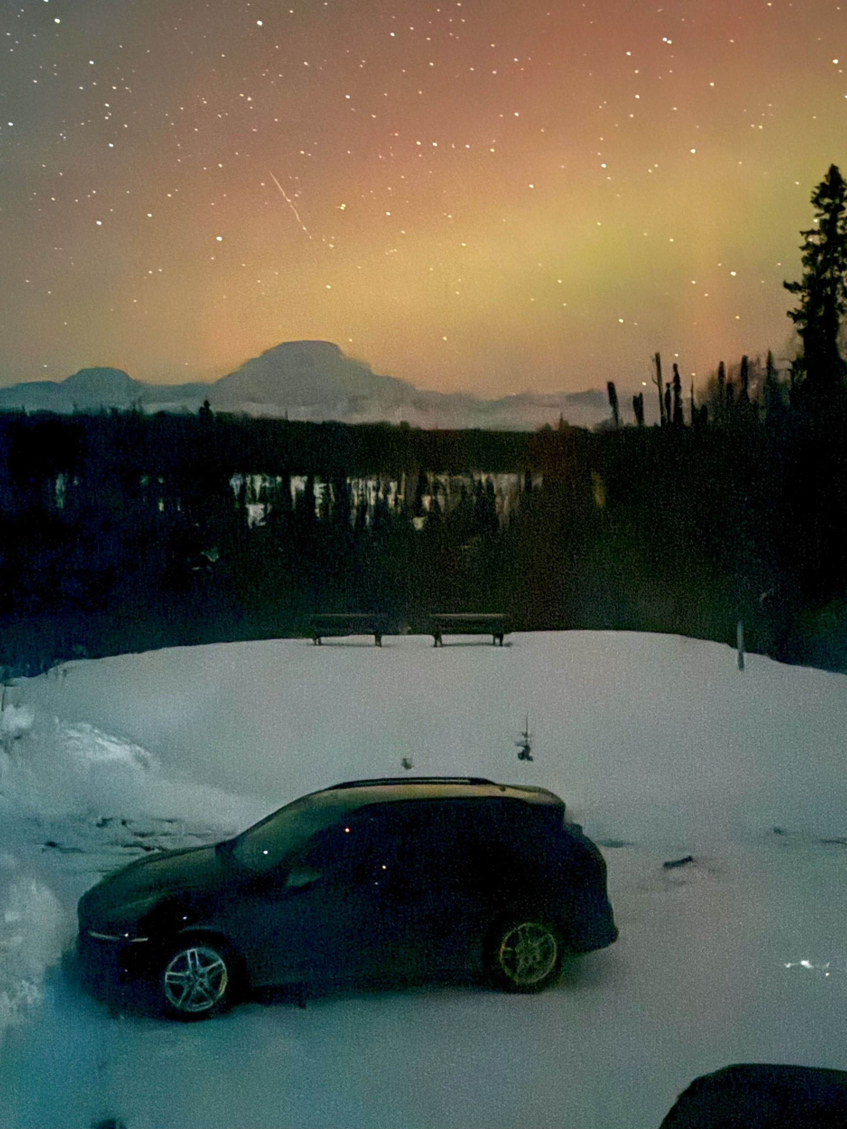 Un coche aparcado en una ladera nevada, con una montaña al fondo y un cielo estrellado arriba.