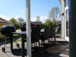 Eine überdachte Terrasse mit Grill, Gartenmöbeln und Blick auf Häuser im Hintergrund.
