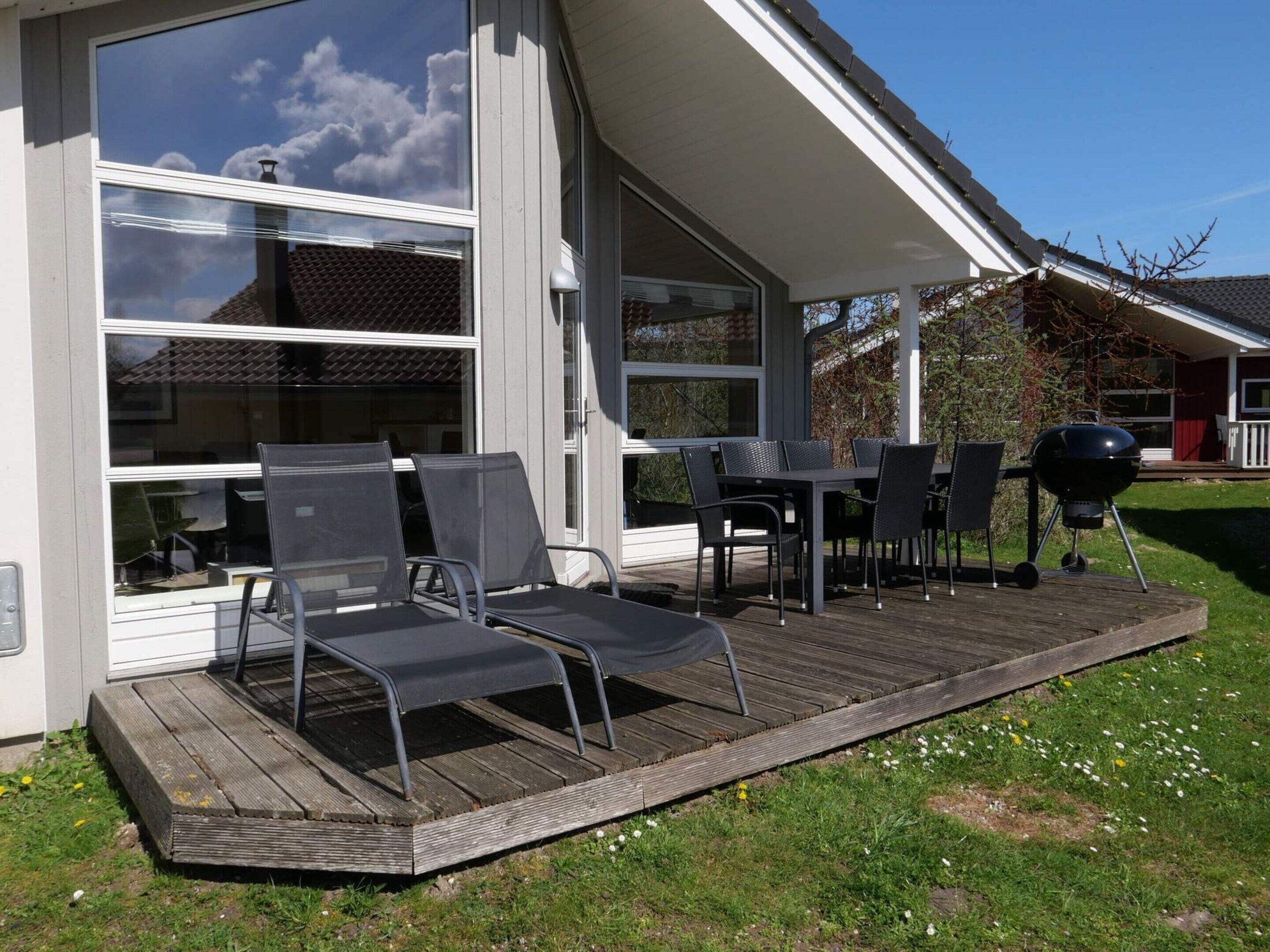 Un patio techado al aire libre con una mesa, sillas, una parrilla y vistas a una casa con ventanas grandes.