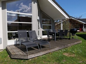 Eine überdachte Terrasse im Freien mit Tisch und Stühlen, einem Grill und Blick auf ein Haus mit großen Fenstern.