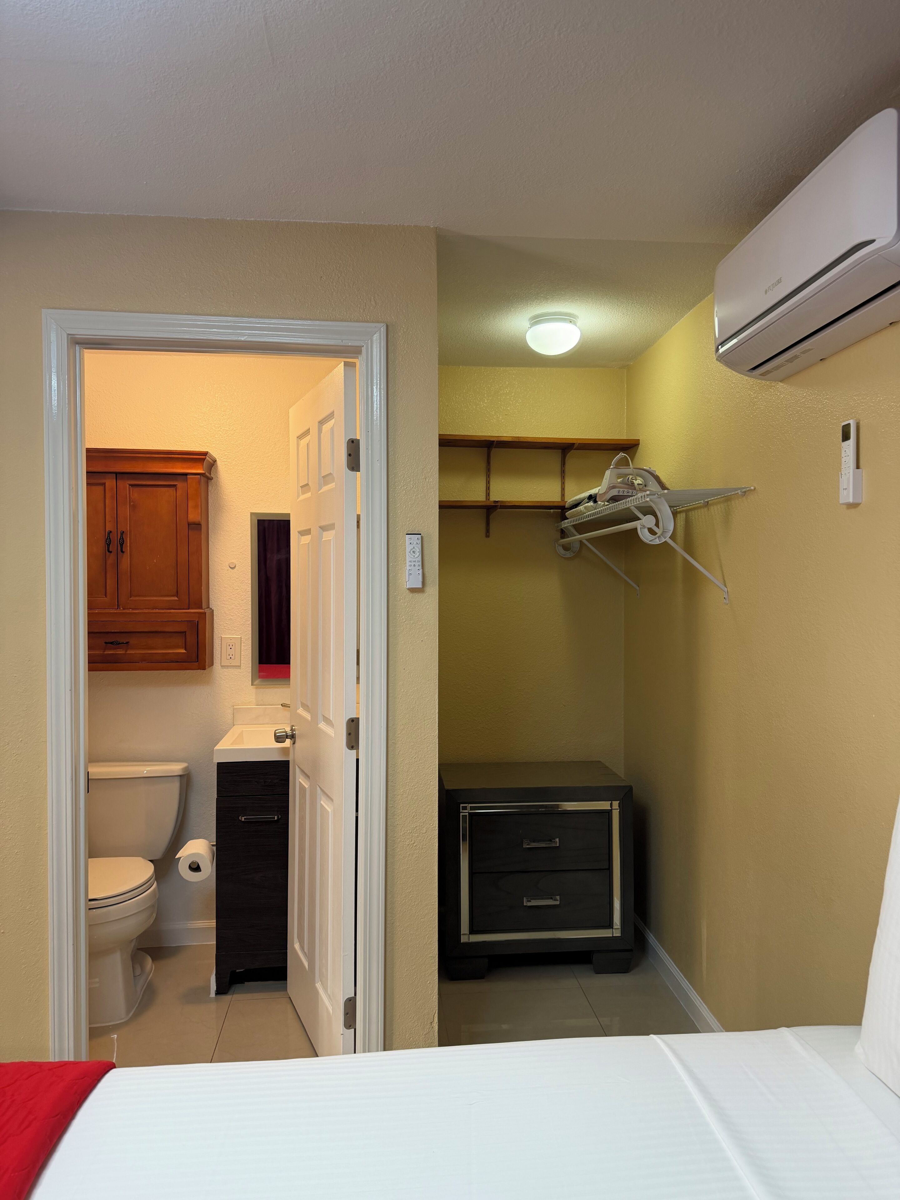 Ein Schlafzimmer mit Bett, Nachttisch, Kleiderschrank und einem Badezimmer inklusive Toilette sowie einer Klimaanlage.