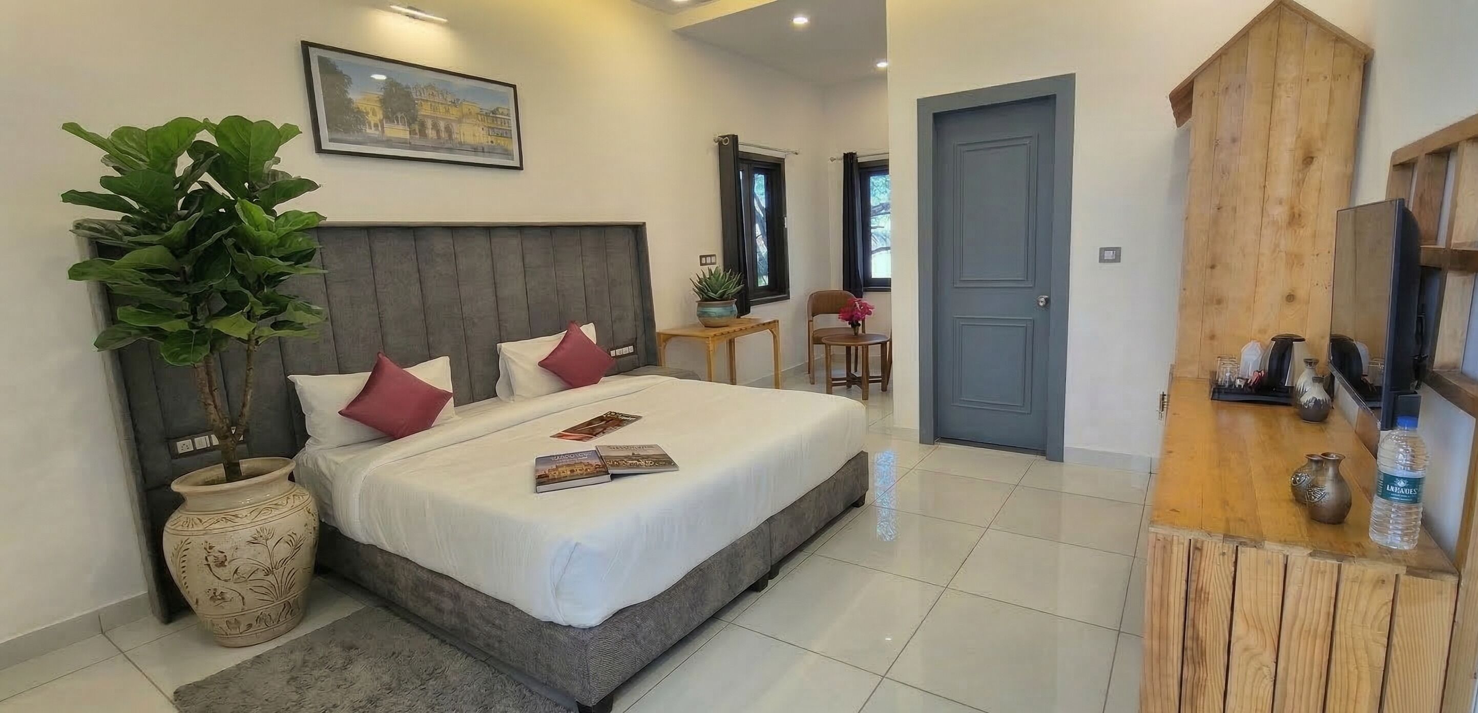 A bedroom with a bed, a potted plant, a framed picture, a small table with a chair, and a wooden reception desk.