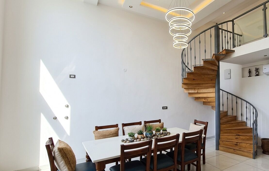 Un coin repas avec une table et des chaises blanches, un escalier avec des marches en bois et un luminaire moderne.