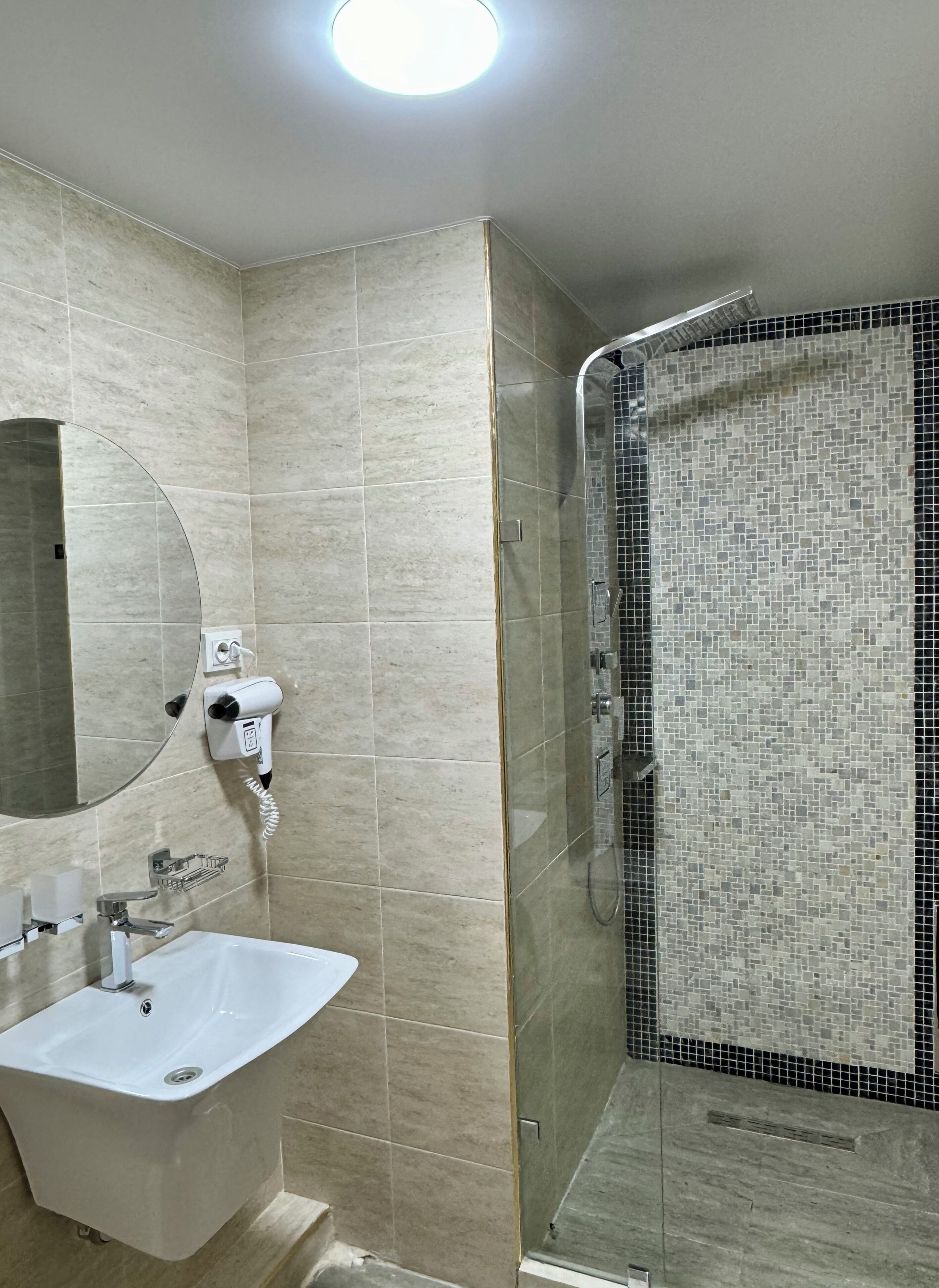 Un baño con un lavamanos blanco, un espejo, un secador de pelo y una ducha con mosaicos.