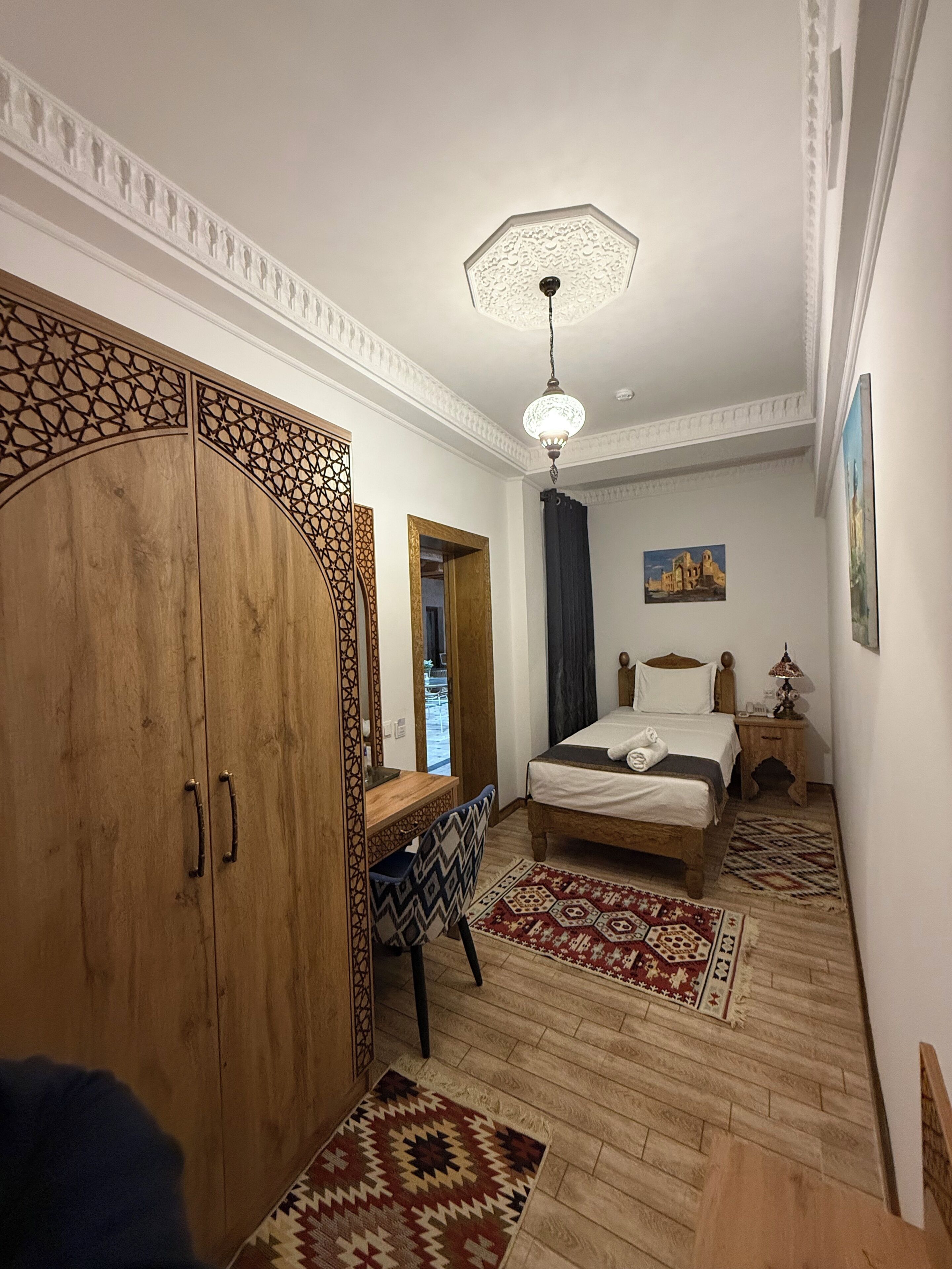Un dormitorio con un armario de madera, una cama con cabecero, un escritorio, una silla, una alfombra estampada y una lámpara en el techo.