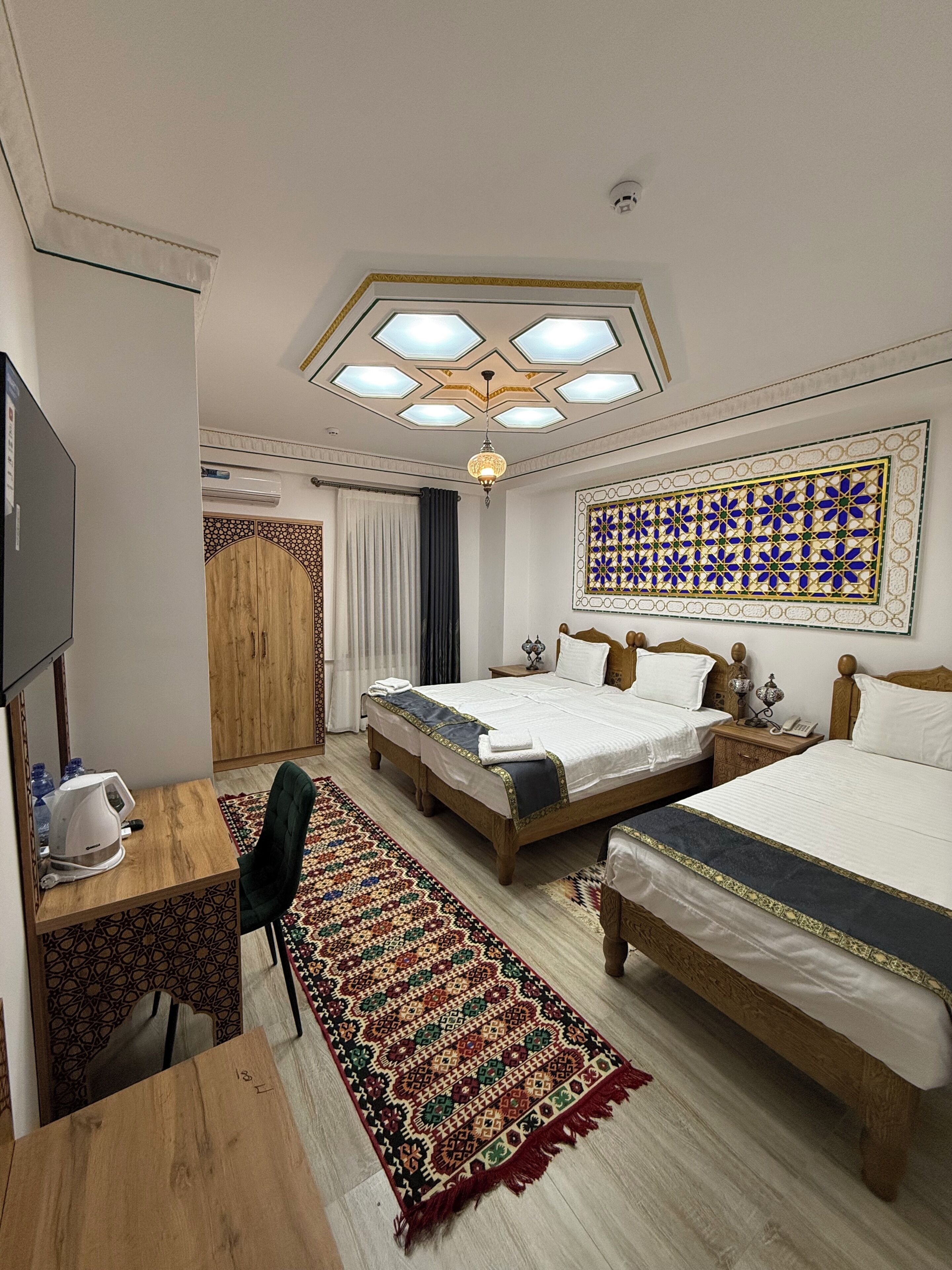 A hotel room with two beds, a desk with a kettle, a TV, and a patterned rug.