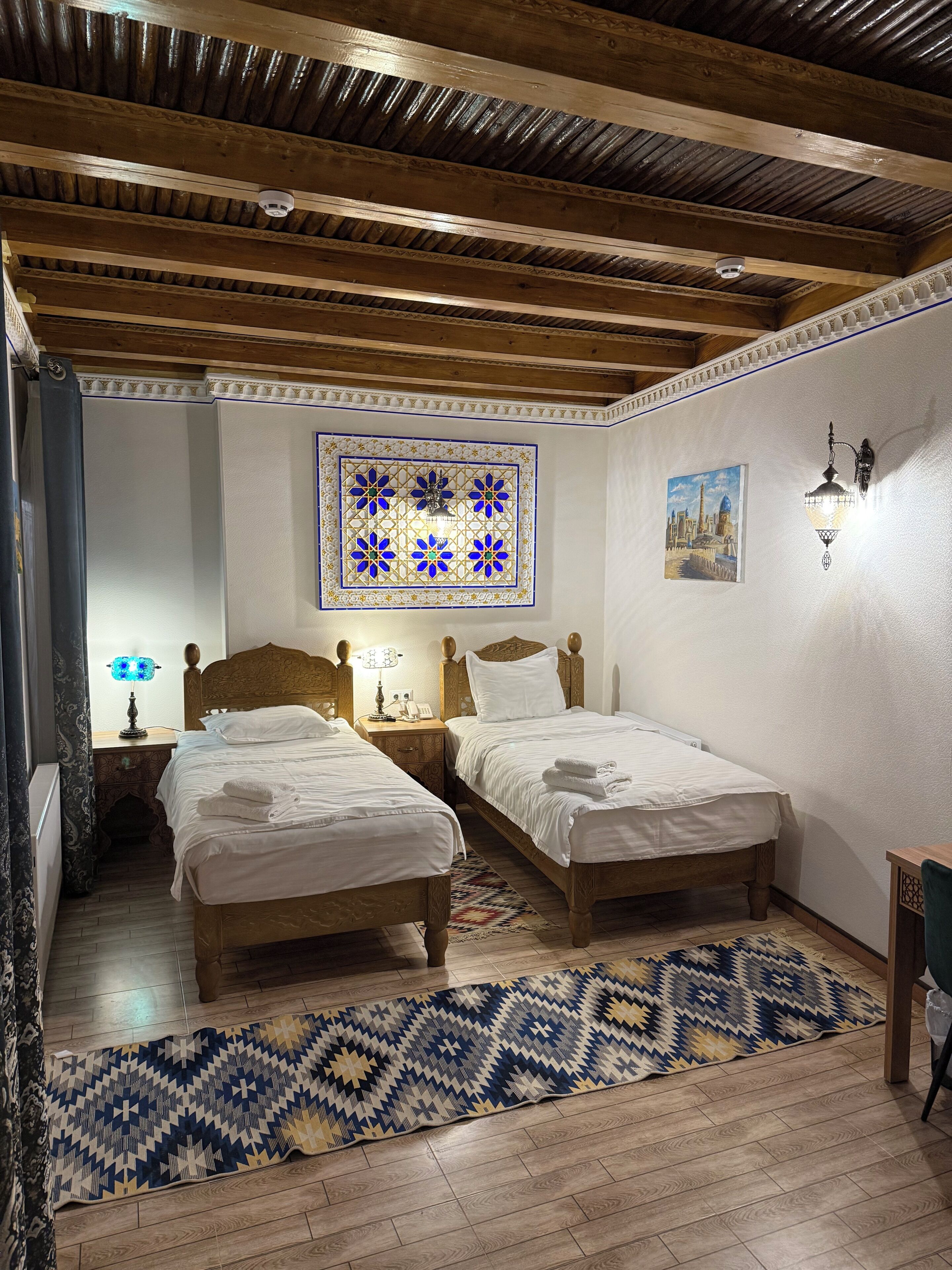 Un dormitorio con dos camas de madera, un tapete estampado, un adorno en la pared y una mesita de noche con lámpara.